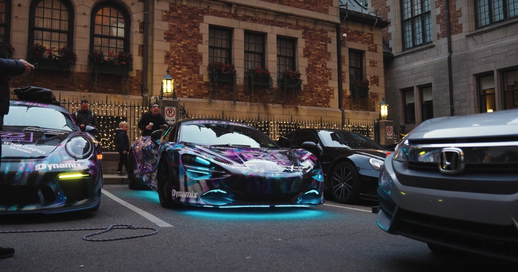 A lineup of high-end rally cars, including a neon-lit McLaren and a Porsche with custom vinyl wraps, parked on a city street in front of a historic brick building decorated with string lights and flower boxes. People stand nearby, including a child admiring the vehicles.