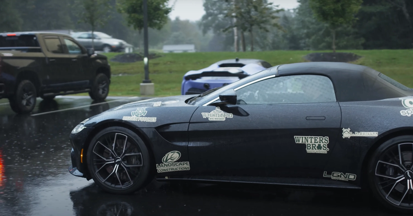 A dark car exists a parking lot with a green grassy field behind it while it resides on a darkened pavement from rain. The car is a black sports car, detailed with branding for a car rally for R2G. There are other trucks in the image as well, escaping the rain in an epic array.