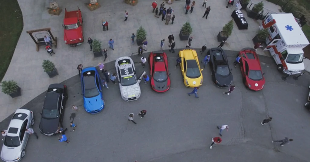 A line of multicolored cars with scattered people all around from the view from the clouds for this Car Rally send off.