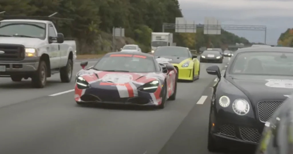 A group of high-end sports cars, including a McLaren with a red, white, and blue rally wrap, drive on a highway during the Rally 2 Give event, surrounded by other rally and regular vehicles.