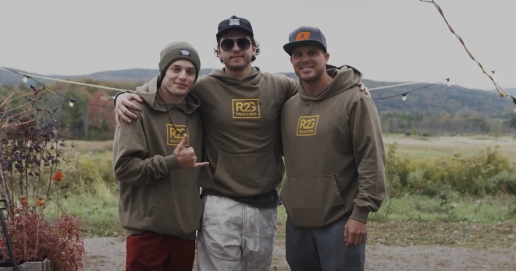 Three men wearing matching olive green 'R2G Rally 2 Give' hoodies pose together outdoors with mountains and fields in the background, under a string of hanging lights.
