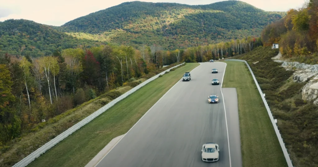A line of cars driving on a racetrack surrounded by autumn-colored trees and rolling forested hills in the background.