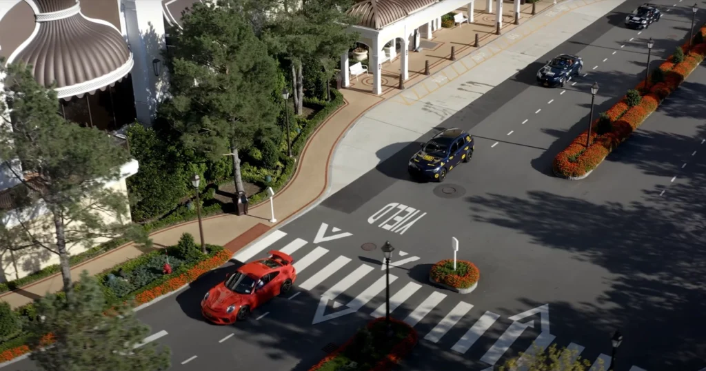 Overhead view of a convoy of rally cars, including a bright red Porsche and several dark-colored vehicles with event decals, driving past landscaped medians and a white portico entrance surrounded by trees and flower beds