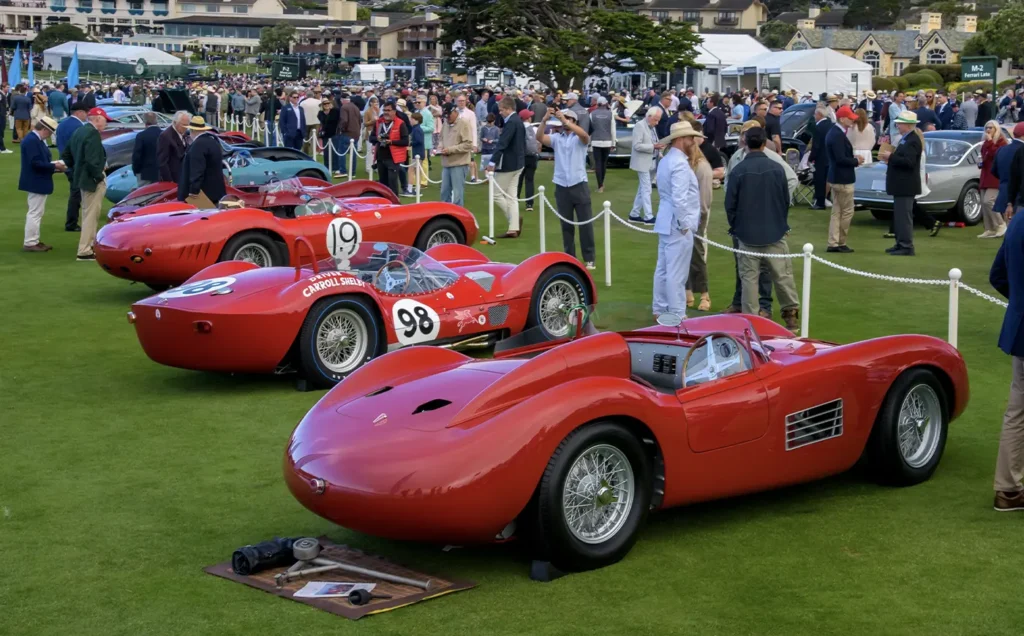 Red vintage race cars on display at the Audrain Newport Concours d’Elegance, with a crowd of spectators gathered on a sunny day.